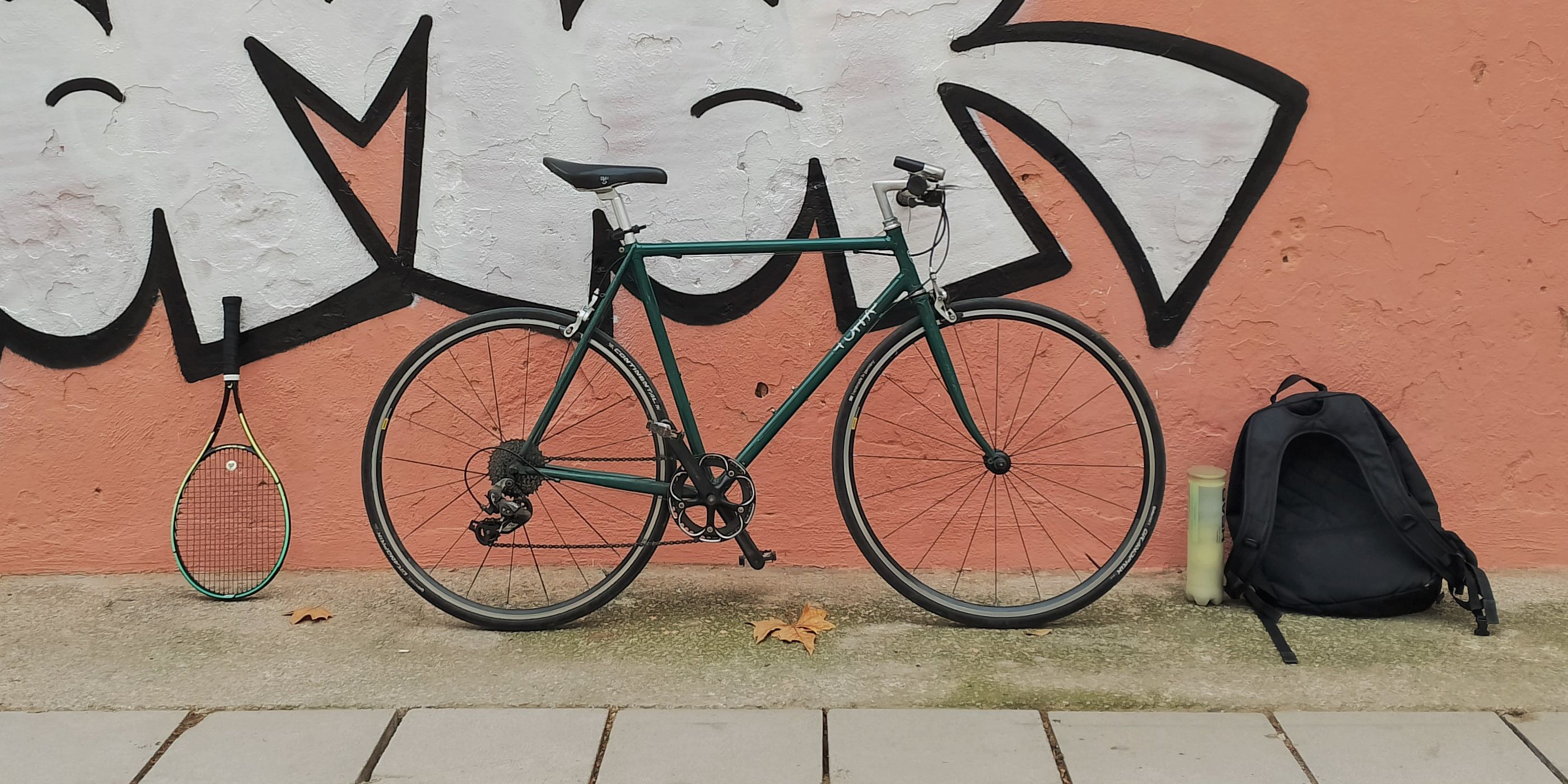 Photo of a city bike, a backpack, and a tennis racquet standing against a graffitied wall.