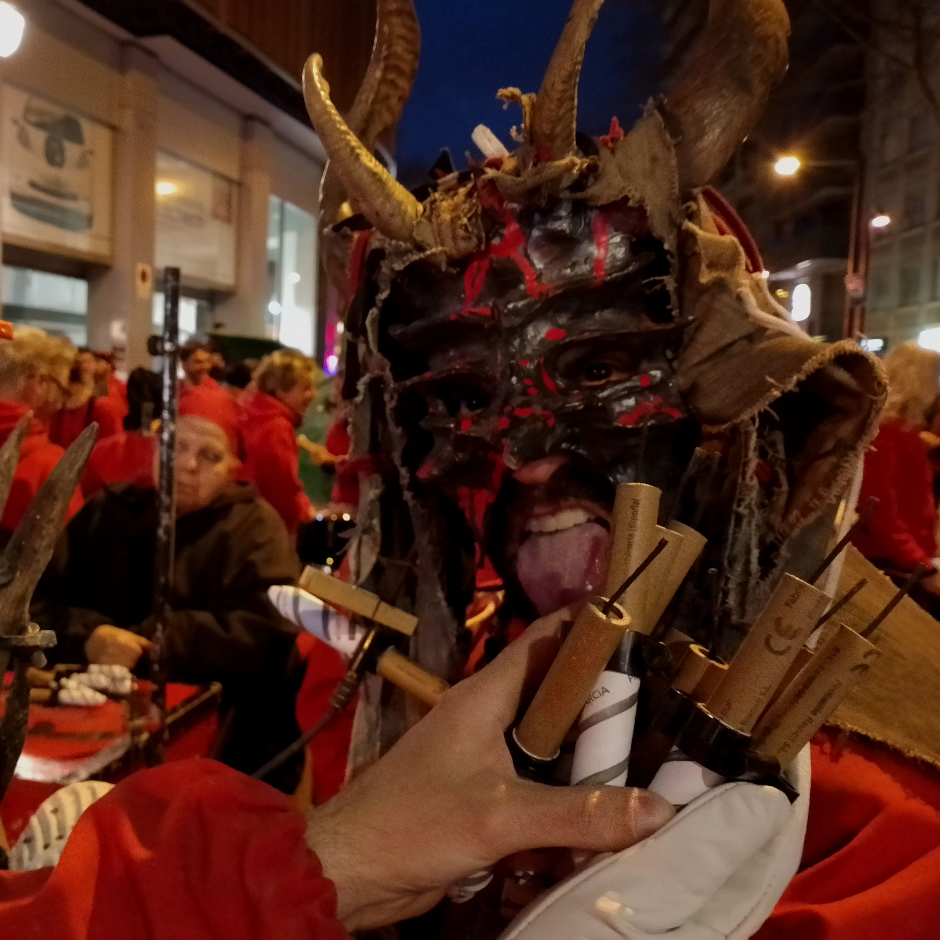 Photo of a diable dressed with a horned hoodie, sticking his tongue out while a hands holds several explosive cartridges next to his face.