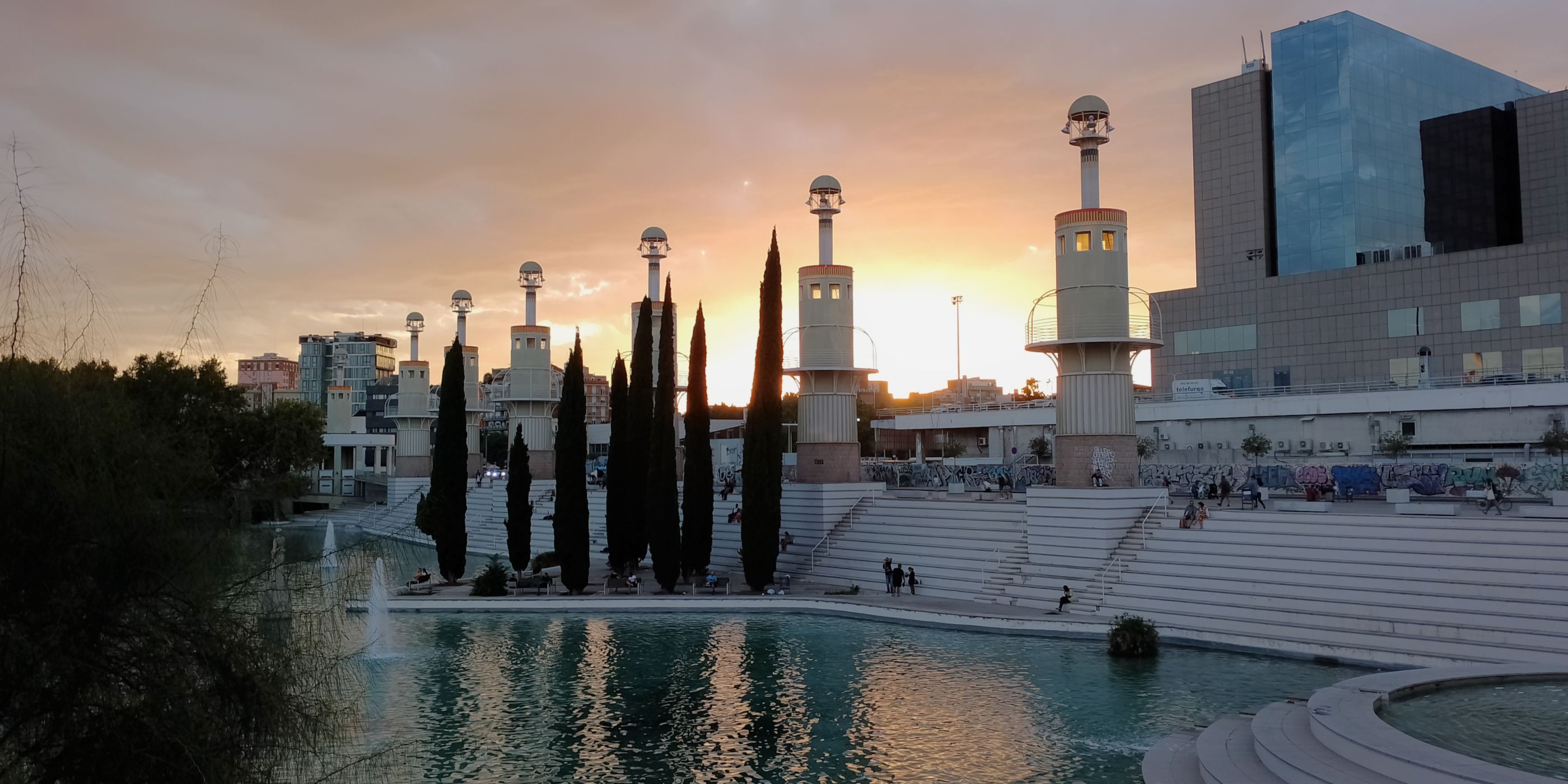 Photo of a park in Barcelona at sunset. There is a shallow green lake on the foreground a series of towers that look like lighthouses, a modern building in the background