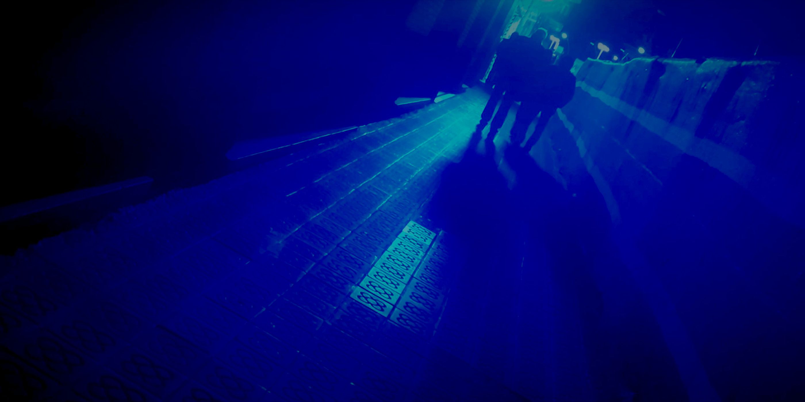 Photo of a sidewalk taken at night, with low light. There is a strong blue light in the background, and everything is tinted blue. The photo is tilted, there are two figures walking but we can only see their dark silhouette.