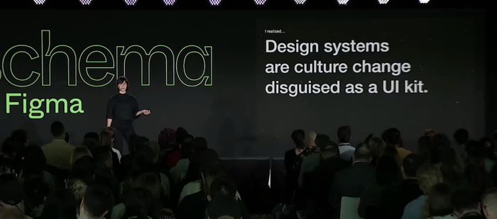 Photo of Lauren LoPrete on stage during Schema 2022, in front of a slide that reads: Design Systems are culture change disguised as a UI kit