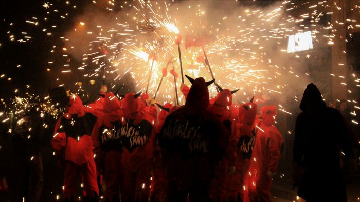 Photo of a dozen people dressed in a red devil suit, with hoodies, black horns, a black bat drawn over their backs and the words Diables de Sants. They are all holding up powerful sparkling sticks, like a meter above their heads, and not like your regular birthday cake sparkles, more like the power of a thousand solar flares.