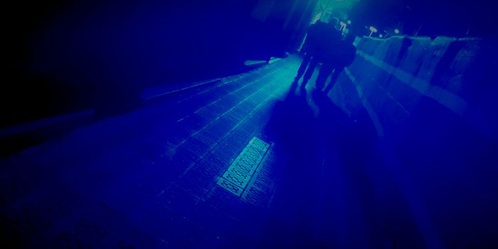 Photo of a sidewalk taken at night, with low light. There is a strong blue light in the background, and everything is tinted blue. The photo is tilted, there are two figures walking but we can only see their dark silhouette.